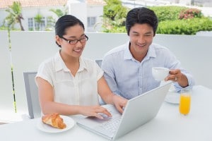 Smiling couple having breakfast together using laptop outside on a balcony